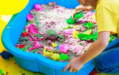 Bir çocuk kinetik kumda kürekle kazar. Fotoğrafta, çocuğun elinde kinetik kumdan yapılmış kum ve plastik oyuncaklar var. Yüksek kalite fotoğraf