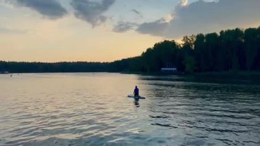 Genç bir adam gün batımında SUP tahtasıyla sakin bir yerden geçiyor. Güzel altın bir gün batımı. Doğada bir insanın aktif olarak yeniden yaratılması. Yüksek kaliteli HD görüntüler