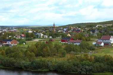 Scenic view of Kautokeino, a small town in Finnmark, Norway, showcasing traditional Nordic houses, a prominent church spire, and lush greenery under a partly cloudy sky clipart