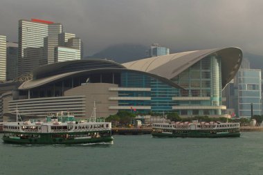 The Hong Kong Convention and Exhibition Centre with its modern curved architecture, set against a skyline of glass skyscrapers, with two Star Ferries crossing Victoria Harbour under a cloudy sky. clipart