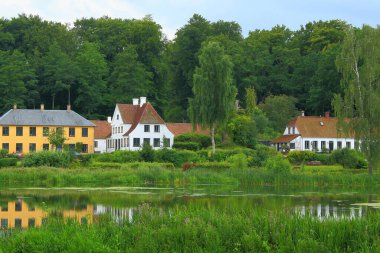 Kırmızı ve sarı cepheli büyüleyici kır evleri yemyeşil sularla çevrili sakin bir gölete yansıyor. Geleneksel mimarinin ve doğanın güzelliğinin sergilendiği sakin kırsal bir ortam.