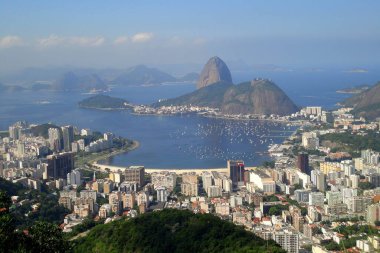 Rio de Janeiro 'nun panoramik manzarası. Sugarloaf Dağı, Guanabara Körfezi ve açık mavi gökyüzü altındaki yemyeşil tepelerle çevrili şehir manzarası.