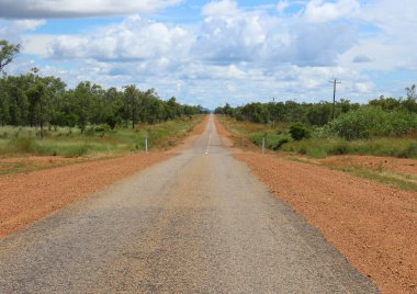 Kırsal kesimde düz bir yol, Avustralya çalılıklarından geçiyor. Dalgalı mavi bir gökyüzünün altında, dağınık bulutlarla. Kuzey bölgesindeki uzak ve doğal güzellikleri gözler önüne seren bir taşra manzarası.