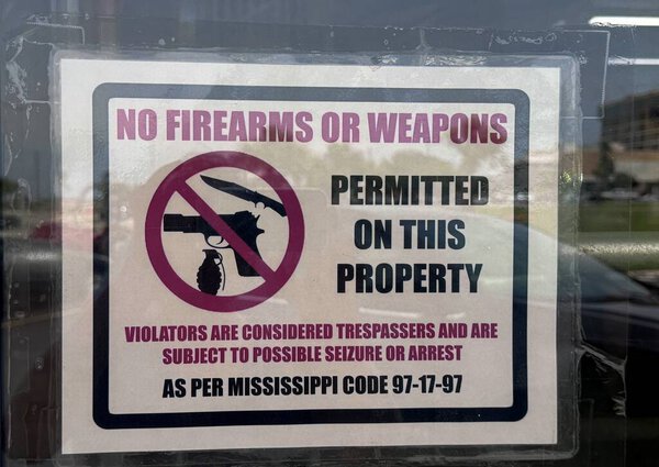 Sign posted on a building prohibiting firearms or weapons on the property, referencing Mississippi Code 97-17-97, with warnings for trespassers and possible arrest or seizure