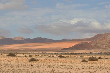 Namib Çölü, Namibya 'da kayan kum tepeleri ve engebeli dağlar, kurak araziye sıcak bir ışık saçıyor, engin ve uzak yabanı aydınlatıyor.