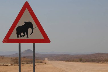 Namibya 'da tozlu çakıllı bir yolda siyah fil siluetinin yer aldığı üçgen kırmızı ve beyaz uyarı levhası sürücüleri olası çöl filleri hakkında uyarıyor.