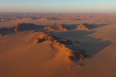 Namibya 'nın Namib Çölü' ndeki engebeli dağ tepeleri ve kumulların hava manzarası, güneş doğarken sıcak hava balonundan yakalanmış, çöl manzarası boyunca uzanan uzun gölgeler.