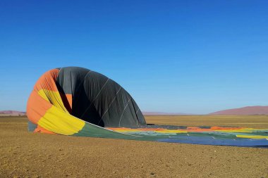 Namib Çölü, Namibya 'ya indikten sonra sönmüş bir sıcak hava balonu çorak çöl zemininde duruyor. Parlak kumaş, berrak mavi gökyüzünün altındaki uçsuz bucaksız, kurak manzarayla çelişiyor.