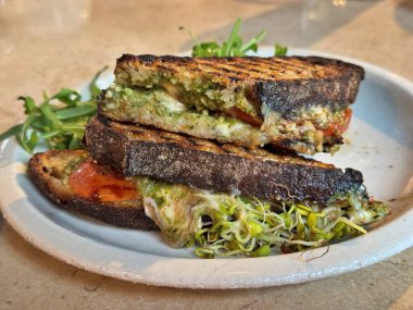 Eritilmiş mozzarella, tavuk, pesto, domates dilimleri ve lahanayla dolu ızgara ekşi ekmek, seramik tabakta sıcak doğal ışıklandırmada roka garnitürle servis ediliyor.