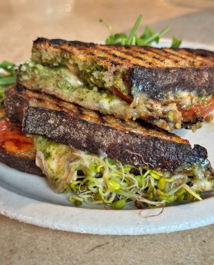 Eritilmiş mozzarella, tavuk, pesto, domates dilimleri ve lahanayla dolu ızgara ekşi ekmek, seramik tabakta sıcak doğal ışıklandırmada roka garnitürle servis ediliyor.