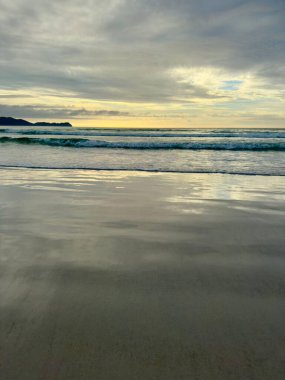 Sakin bir sahil üzerinde sakin bir gün doğumu, yumuşak dalgalar bulutlu bir gökyüzünün altında pürüzsüz kumları emiyor, huzurlu ve sakin bir sabah manzarası yaratıyor..