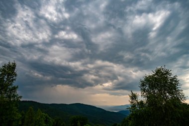 Görkemli bir dağ sırasının üzerinde beliren bulutlu bir gökyüzü, bereketli ağaçlarla dolu sakin bir ön planla tamamlanmış.
