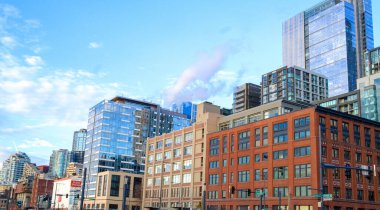 Parlak, berrak bir mavi gökyüzü altında, hayat dolu bir kentsel çevrenin resmedildiği geniş bir dizi binanın bulunduğu canlı bir şehir manzarası.