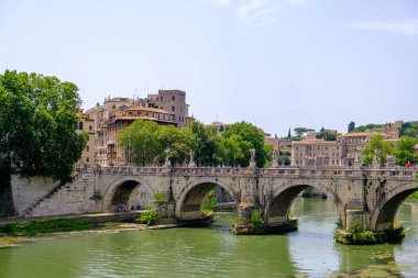 Huzurlu bir nehir boyunca uzanan resimsel bir köprü, arka planda güzel bir şekilde yerleştirilmiş binalar ve yapılar.