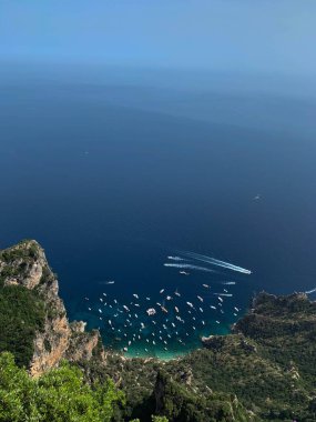 İtalya 'daki Capri Adası' nın panoramik görüntüsü, yüksek görüş açısına sahip. Görüntü, çok sayıda tekne ve yatla derin mavi Akdeniz 'i gözler önüne seriyor. Capri 'nin engebeli kıyı şeridinde dramatik uçurumlar ve yemyeşil alanlar var.,
