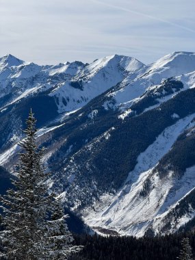 Fotoğraf, karlı bir dağ sırasının yakın görüntüsünü yakalıyor, engebeli zirveleri ve karla kaplı arazinin karmaşık detaylarını gösteriyor. Sahne, açık mavi gökyüzünün altındaki dağların güzelliğini ve görkemini vurgular..