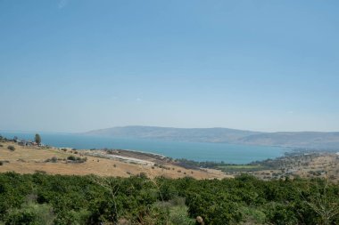 Fotoğraf, İsrail 'deki Güzellik Dağı' ndan görülen Celile Denizi 'nin panoramik bir görüntüsünü yakalar. Sahne, bereketli yeşillikler ve yuvarlanan tepelerle çevrili gölün sakin mavi sularını içeriyor. Huzurlu ve tarihi seti yansıtıyor.