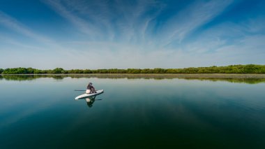 Berrak bir gökyüzünün altında, sakin ve yansıtıcı sularda, yemyeşil sularla çevrili, sükunet, keşif ve doğa ile bir bağlantı içeren sakin bir kürek çekme deneyimi..