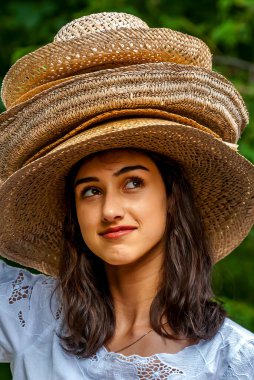 Genç bayan sıcacık gülümsüyor ve birkaç hasır şapka takıyor, açık havada yemyeşil, neşeli ve neşeli bir hava sergiliyor..