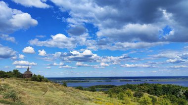 Tepeli yaz manzarası ve geniş bir nehre bakan geleneksel ahşap kilise kümülüs bulutlu mavi gökyüzünün altında.