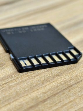 Ahşap bir yüzeyde duran mikro SD kartın detaylı bir görüntüsü. Gösterişli siyah tasarım, birden fazla aygıt arasında veri depolamak için kullanılan temel teknolojiyi vurgulayan altın tonlu temas noktalarını içeriyor. Modern ürünleri sergilemek için ideal