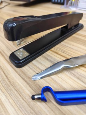 Siyah metal zımbanın yakın plan görüntüsü ahşap bir masaya yerleştirilmiş, yanında parlak gümüş bir mektup açacağı ve ortak ofis çalışma ortamını temsil eden mavi bir stilus kalem var..