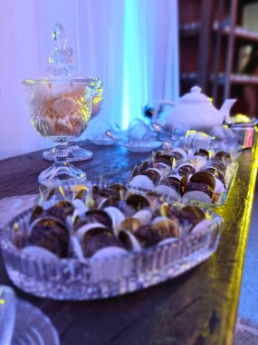 Yumuşak ortam ışıklarıyla aydınlatılan çeşitli şekerlerle dolu kristal tabaklarla zarif bir şekilde düzenlenmiş bir tatlı masası. Çaydanlık ve şeker kasesi sofistike ortamı tamamlar..