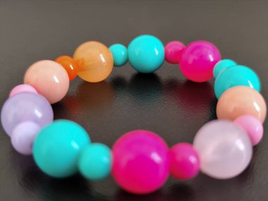 Pembe, mavi, turuncu ve mor tonlarda renkli plastik boncuklardan yapılmış canlı bir bilezik, düz siyah bir arkaplanda duruyor..