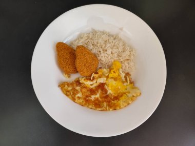 Beyaz tabakta sunulan lezzetli bir yemek. Tabakta bol pirinç, altın omlet ve kızarmış tavuk nugget var..