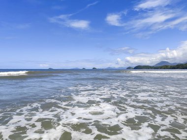 Güneşli bir günde, yumuşak dalgaların kuma vurduğu sakin bir deniz manzarası. Açık mavi gökyüzü, arka planda suya yansıyan küçük adalarla sakin manzarayı tamamlar..