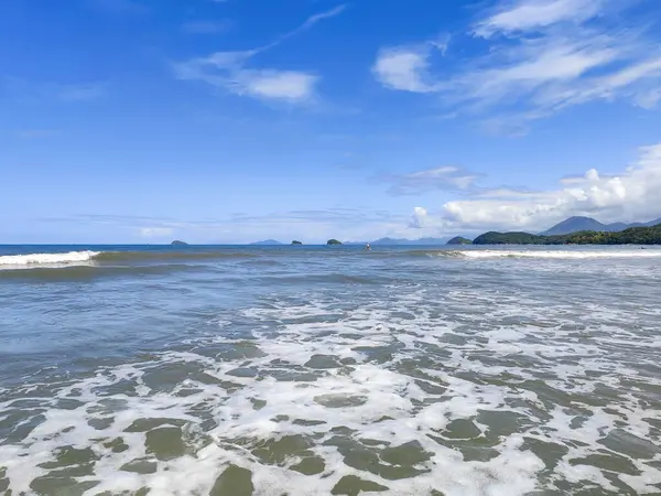 Güneşli bir günde, yumuşak dalgaların kuma vurduğu sakin bir deniz manzarası. Açık mavi gökyüzü, arka planda suya yansıyan küçük adalarla sakin manzarayı tamamlar..