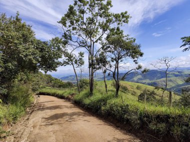 Bereketli beyaz bulutlarla dolu açık mavi bir gökyüzünün altında yeşil bir dağ manzarası boyunca uzanan toprak bir patika. Yol kenarındaki ağaçlar manzaranın sükunetini tamamlıyor. Cunha SP