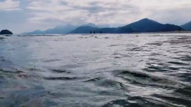 Plajlar arasındaki sığ okyanus suyunda yürüyen insanların huzurlu manzarası, yumuşak ışık, bulutlu gökyüzü ve arka planda yemyeşil dağlar..