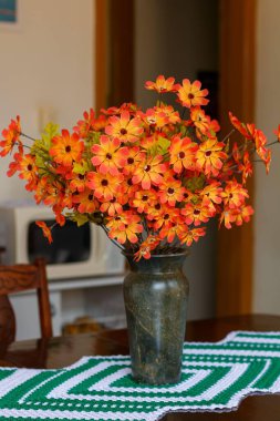 A beautiful arrangement of orange flowers in a sturdy green, marble-like vase, placed on a wooden table with a decorative knitted table runner.  clipart