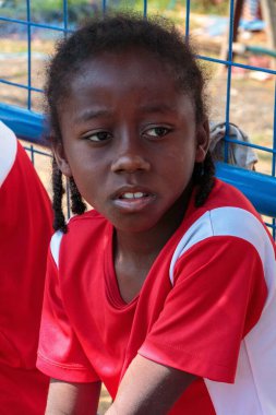 Kırmızı-beyaz formalı Afrikalı genç bir futbolcu maç ya da antrenman sırasında dikkatlice kenarda oturuyor..