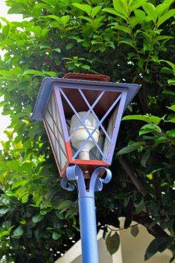Şehir parkında renkli mavi bir vücudu ve kırmızı detayları olan dekoratif bir fener. Arka planda, sık yeşil bir çalı doğanın güzelliğini arttırır..
