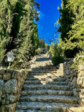 Kırsal taştan bir merdiven, yükselen selvi ağaçları ve canlı yeşilliklerle çevrili, gür bir Akdeniz bahçesinden yukarıya doğru uzanır. Sıcak taş duvarlar ve klasik fenerler sonsuz bir cazibe yaratırken derin mavi gökyüzü renkleri güçlendiriyor..