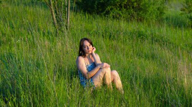 Yazın yeşil bir çayırda oturan uzun siyah saçlı genç bir kadın. Etrafı uzun otlar ve kır çiçekleriyle çevrili. Sıcak altın güneş ışığının tadını çıkarıyor..