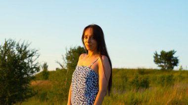 Yazın yeşil bir çayırda oturan uzun siyah saçlı genç bir kadın. Etrafı uzun otlar ve kır çiçekleriyle çevrili. Sıcak altın güneş ışığının tadını çıkarıyor..