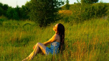 Yazın yeşil bir çayırda oturan uzun siyah saçlı genç bir kadın. Etrafı uzun otlar ve kır çiçekleriyle çevrili. Sıcak altın güneş ışığının tadını çıkarıyor..