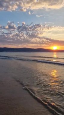 Bulgaristan 'ın Karadeniz kıyı şeridinde canlı bir güneş doğuyor. Güneşli sahil beldesi, şafak sahnesi güneş ufukta yükselen tepeler ve kumları yıkayan köpüklü dalgalar.