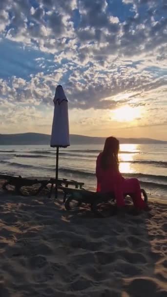 无忧无虑的年轻女子坐在沙滩上的日光浴床上 望着海面上的日出 美丽的海滨黎明美景 旅行和放松的概念 — 图库视频影像