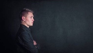 Düşünceli genç adam portresi, kollarını katlanmış bir şekilde arka plan üzerinde fotokopi alanı olan karanlık bir duvara dayıyor. Kararlı yüz ifadesi olan düşünceli bir erkek öğrenci.