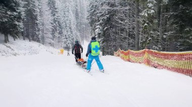 Dağ kurtarma ekipleri yaralı bir kayakçıyı taşıyorlar, ilk yardım sonrası yokuş aşağı gidiyorlar. Yokuş kurtarma, kayak devriyesi özel bir kızakla kurbanı tahliye ediyor. Bukovel kayak merkezi, Ukrayna Karpatları