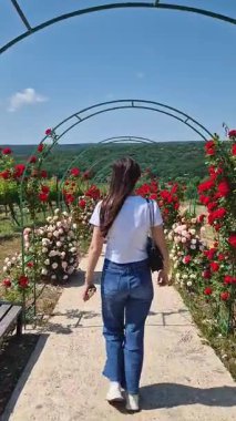 Genç bir kadın, kemerlerdeki çiçeklerle süslenmiş bir gül yolu boyunca yürüyor. Kırmızı ve pembe çiçeklerle çevrili mutlu kız ilerideki kayan tepelere huzurlu bir manzarayla gezintiden zevk alıyor.