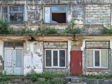 Harabeler içinde terk edilmiş bir bina. Boyası soyulmuş, alçı ve çatlakları parçalanmış. İhmal edilmiş duvarlardan ve yıpranmış kirişlerden fazla büyümüş çalılar ve otlar. Kırık pencereler ve açık tuğlalar çürümeyi gösterir
