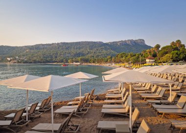 Gündoğumunda Türkiye 'nin Kemer tatil beldesinde Calis Tepesi' nde serene Moonlight plajı. Boş güneş şezlongları ve beyaz şemsiyeler kumlu sahil ve sakin deniz yavaşça kıyıya vurur.