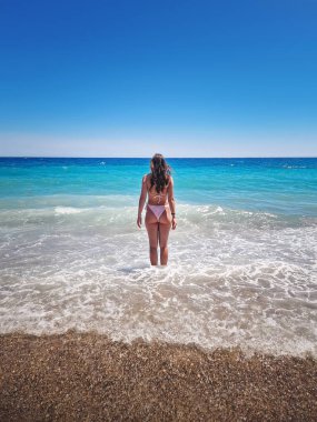 Bikinili güzel bir kadın açık mavi denizin köpüklü dalgalarında duruyor, bulutsuz bir yaz gökyüzünün altında geniş ufka bakıyor. Tropikal bir plajda yaz tatili, rahatlama hisleri