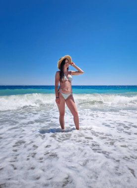 Bikini ve hasır güneş şapkasıyla poz veren güzel bir kadın bulutsuz bir yaz gökyüzünün altında berrak mavi denizin köpüklü dalgalarında duruyor. Tropikal bir plajda yaz tatili, rahatlama hisleri