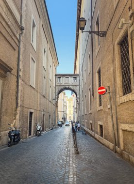 Uzun, dar bir kaldırım taşı caddesi, yüksek tuğlalı binalar ve İtalya 'nın Roma şehrinde eşsiz bir kemer yolu. Park etmiş scooterlar ve şehirde yürüyen birkaç kişi.